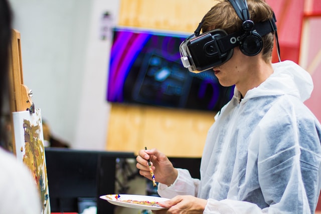 Teenage boy wearing a virtual headset whilst paining a picture in a classroom. He is holding the paintbrush in one hand while looking at the canvas through a the virtual lens.