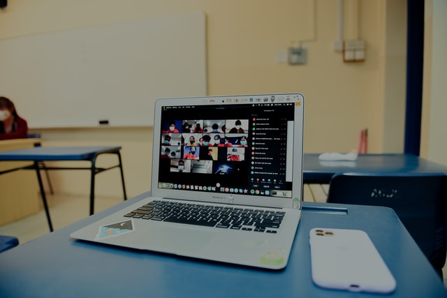 Mac laptop on a school desk showing a video conference call happening while in a classroom. The teacher sits at the front wearing a face mask but the rest of the classroom appears empty.