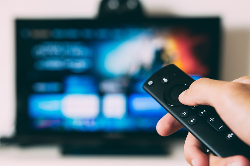 A hand holds a small Firestick remote control in front of a TV. The TV screen is blurred in the background.