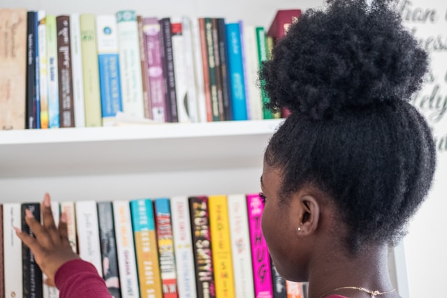 Image seen from behind of a black girl reaches for a book on a book shelf with a colourful selection of book spines.