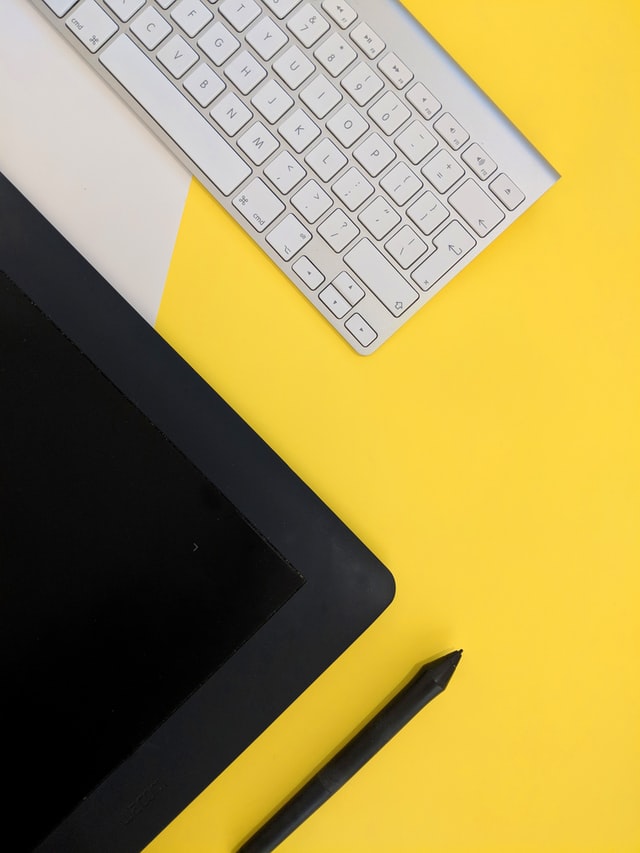 Half of blank laptop screen, a black pencil and a white keyboard above a yellow backdrop.