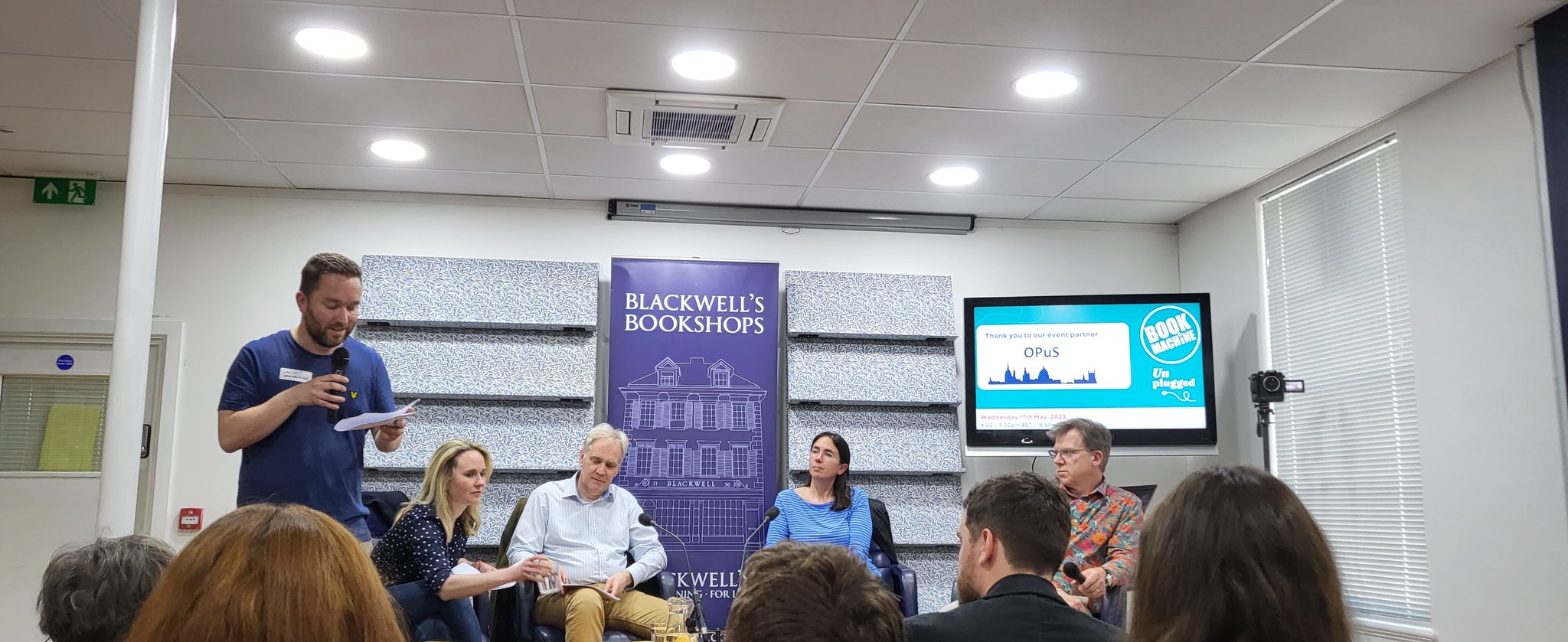 Panel of four adults being introduced by a speaker with a microphone at Blackwell's Bookshop. The image is taken from the audience.